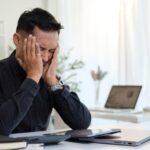 A man in business attire sits at a desk with his head in his hands displaying signs of stress and exhaustion, illustrating what is executive burnout and the toll of workplace pressure.