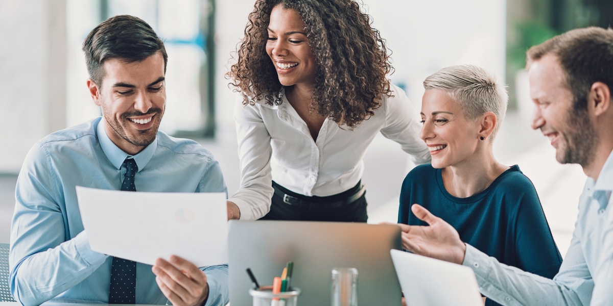 A woman showcasing executive presence for women by confidently engaging her smiling team around a laptop and documents in a bright modern office setting.

