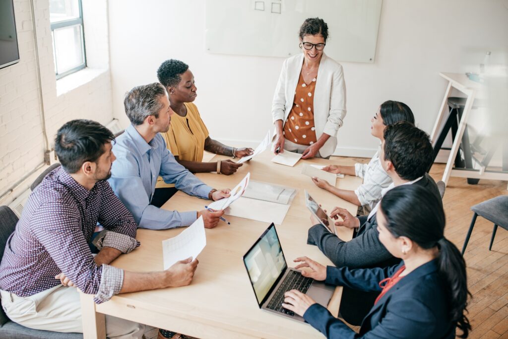 A woman demonstrating executive presence for women by confidently leading a diverse team meeting in a bright modern office while presenting documents to engaged colleagues.